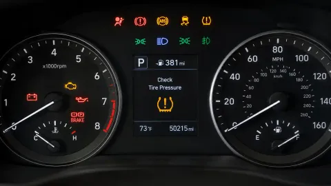 Vehicle instrument cluster illuminated with red, amber, green, and blue dashboard warning lights and a tire-pressure alert.